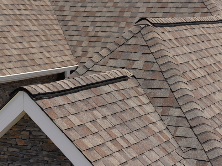 Multi-colored shingles on the gabled roof of a house with stacked stone exterior and white gutters.