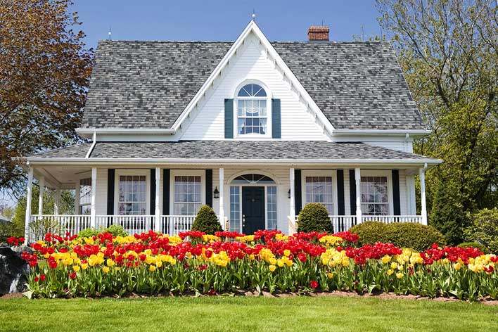White farmhouse-style home with colorful flowers in front White farmhouse-style home with colorful flowers in front