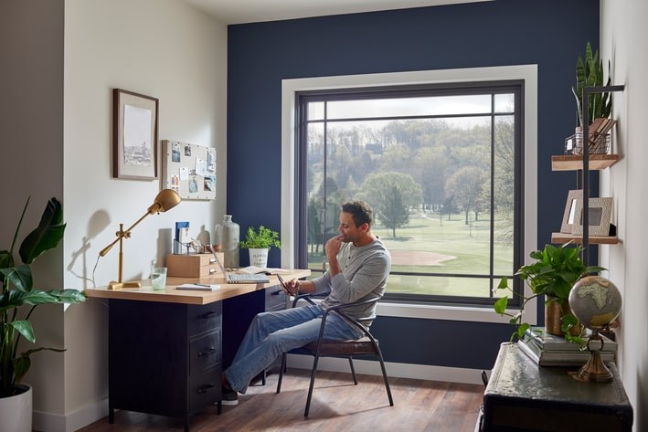 A man looking at his phone while sitting at a desk by a large, square picture window overlooking a golf course.