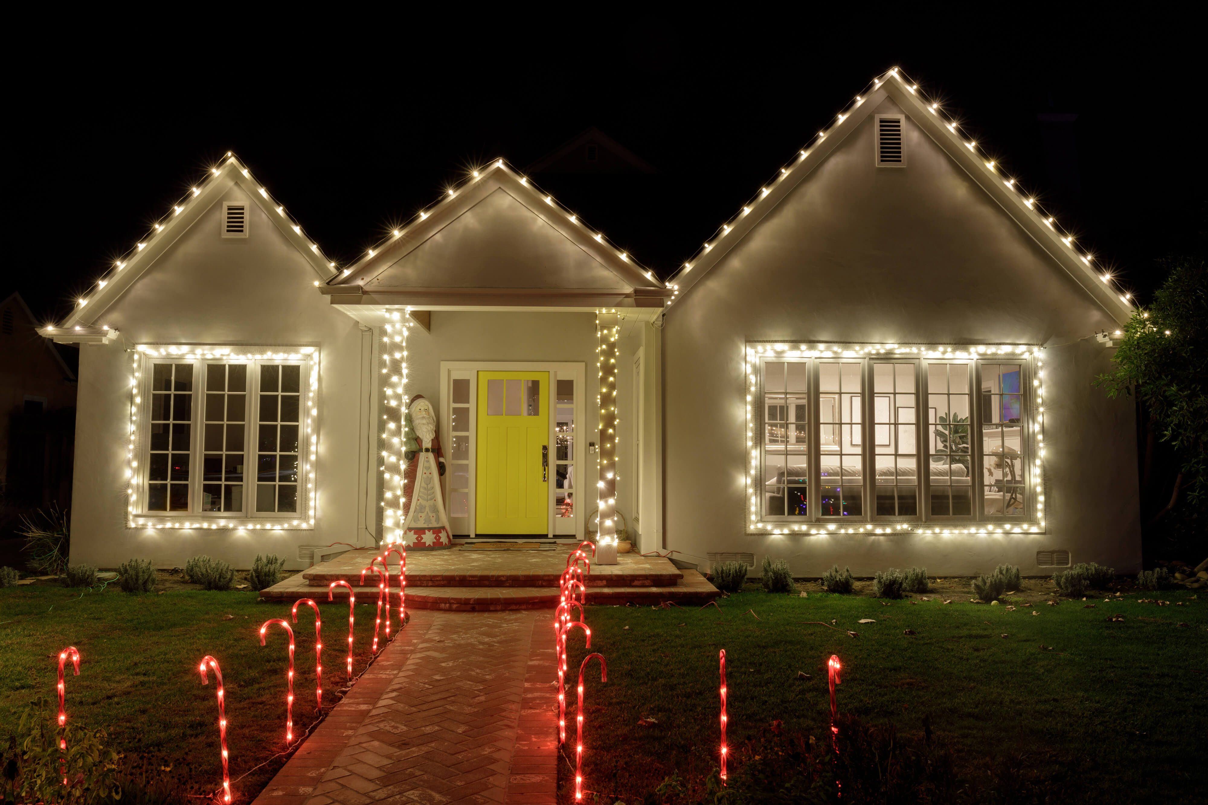 Walkway to a home with a yellow front door and large picture windows decorated with Christmas lights and candy cane lights 