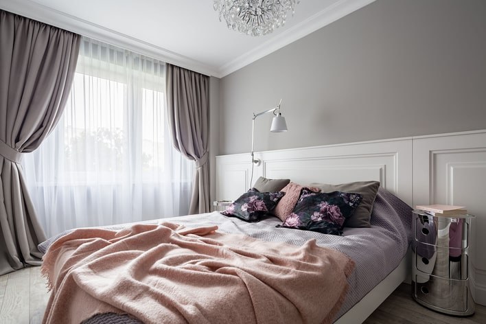 A cozy bedroom with purple and gray tones throughout that has floor-to-ceiling curtains covering the large windows.