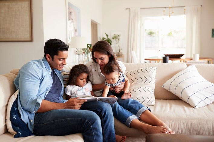 Two parents reading to their two children on the couch in a white, neutrally decorated living room that has large windows covered by sheer curtains.