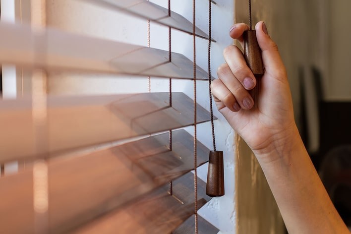 A person pulling the string of wood blinds to close them.