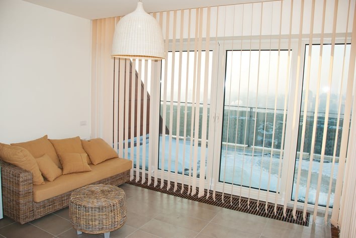 A minimalistic, neutral-toned living room that has a wall of windows that can be covered with vertical blinds.