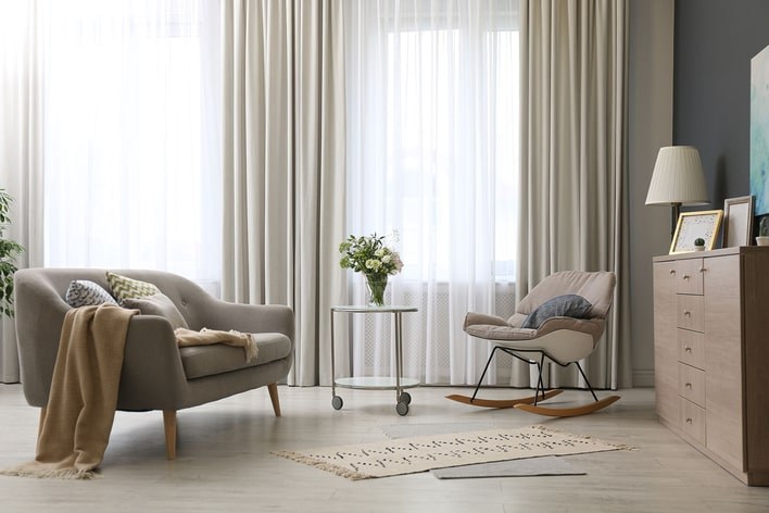 A cozy bedroom nook with neutral-toned furniture that has large windows covered by sheer curtains.