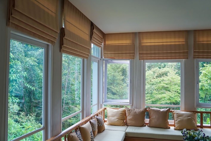 A large, neutral-toned sunroom with walls of large windows that can be covered with Roman shades.