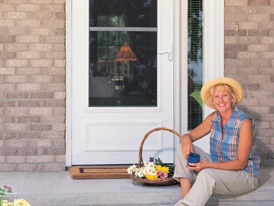 A smiling woman in a yellow hat sits on her porch in front of a ventilating storm door.